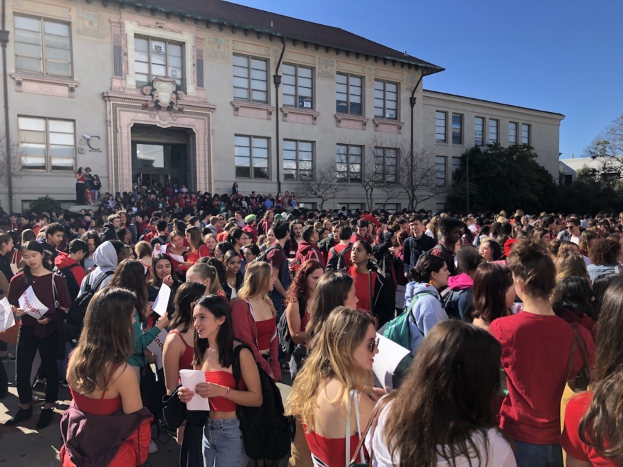 A sea of teenagers outside a school building. Most are wearing red, some are holding pieces of paper.