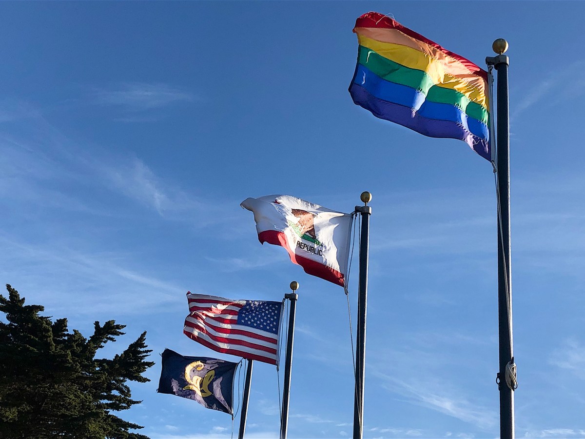 Four flags flying against blue sky including US flag, State of California flag, rainbow flag, and Cal flag