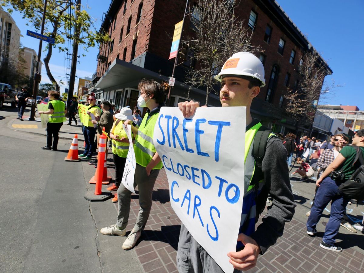 A person in a hard hat and yellow safety vest holds a sign reading "street closed to cars" while standing at the intersection of Telegraph and Durant avenues during a rally near UC Berkeley.